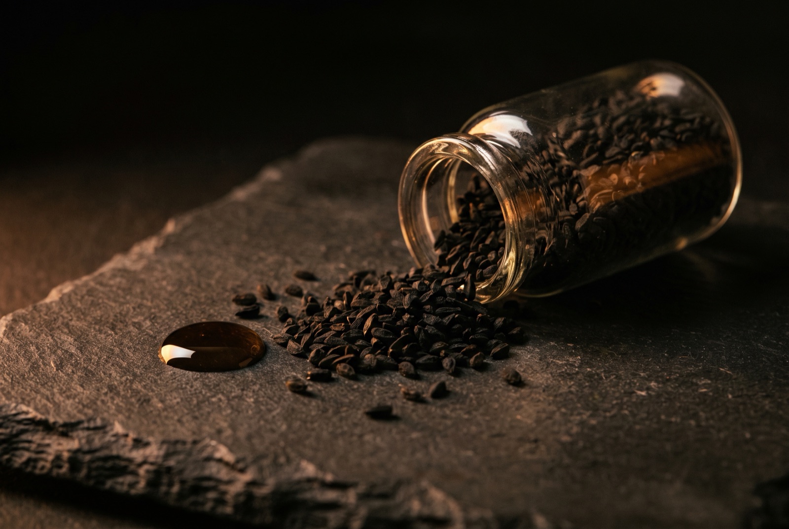 Black cumin seeds spilling from a glass vial beside a drop of dark oil